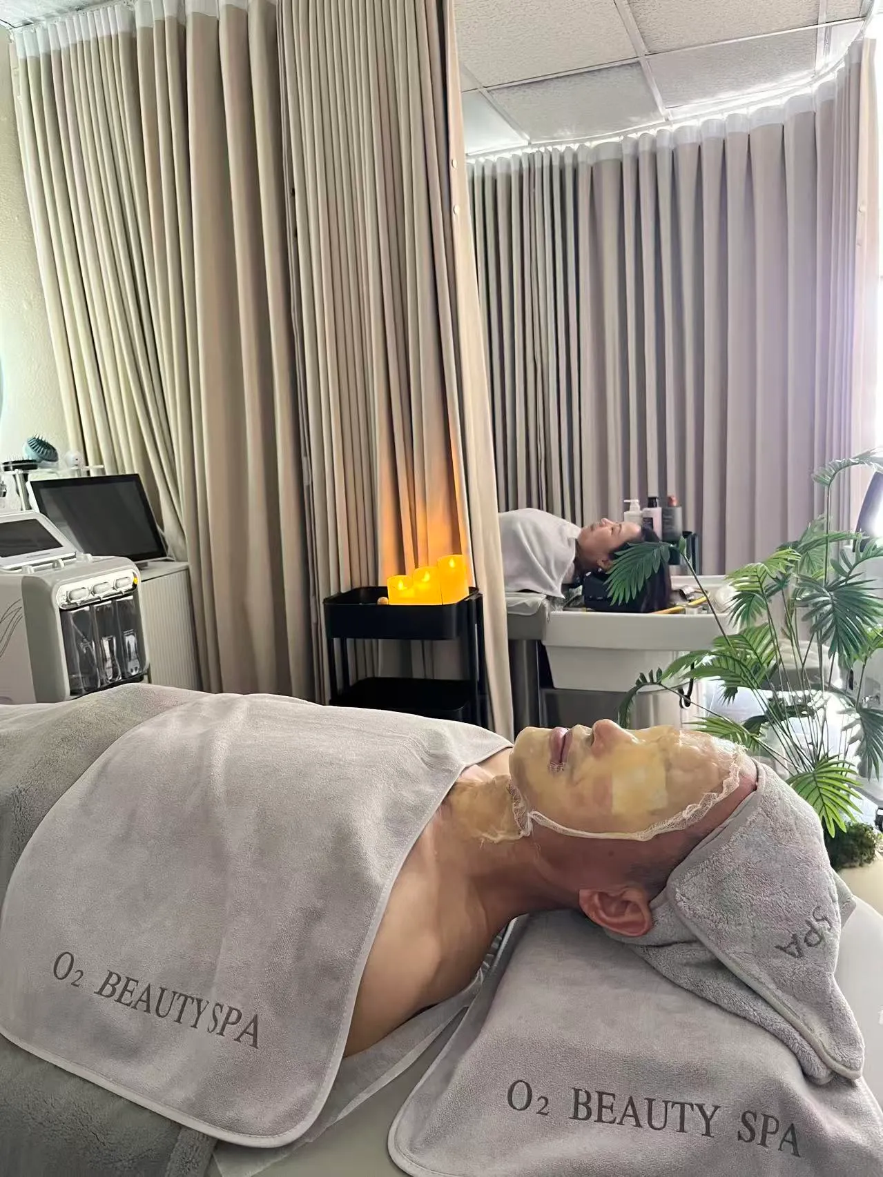 A facial client under O2 Beauty Spa towels in a gold mask, with another client at the head spa basin in the background of the same room