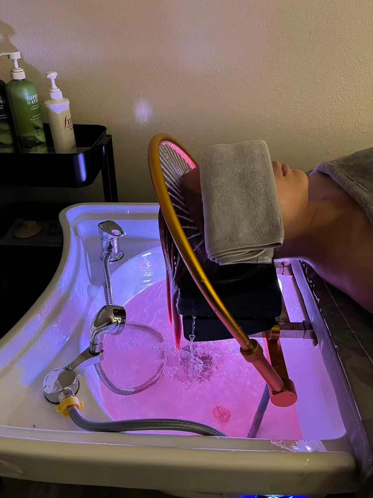 Head spa basin glowing rose-pink at O2 in Arcadia, water running from a gold arch through a client's hair, a folded towel over the eyes