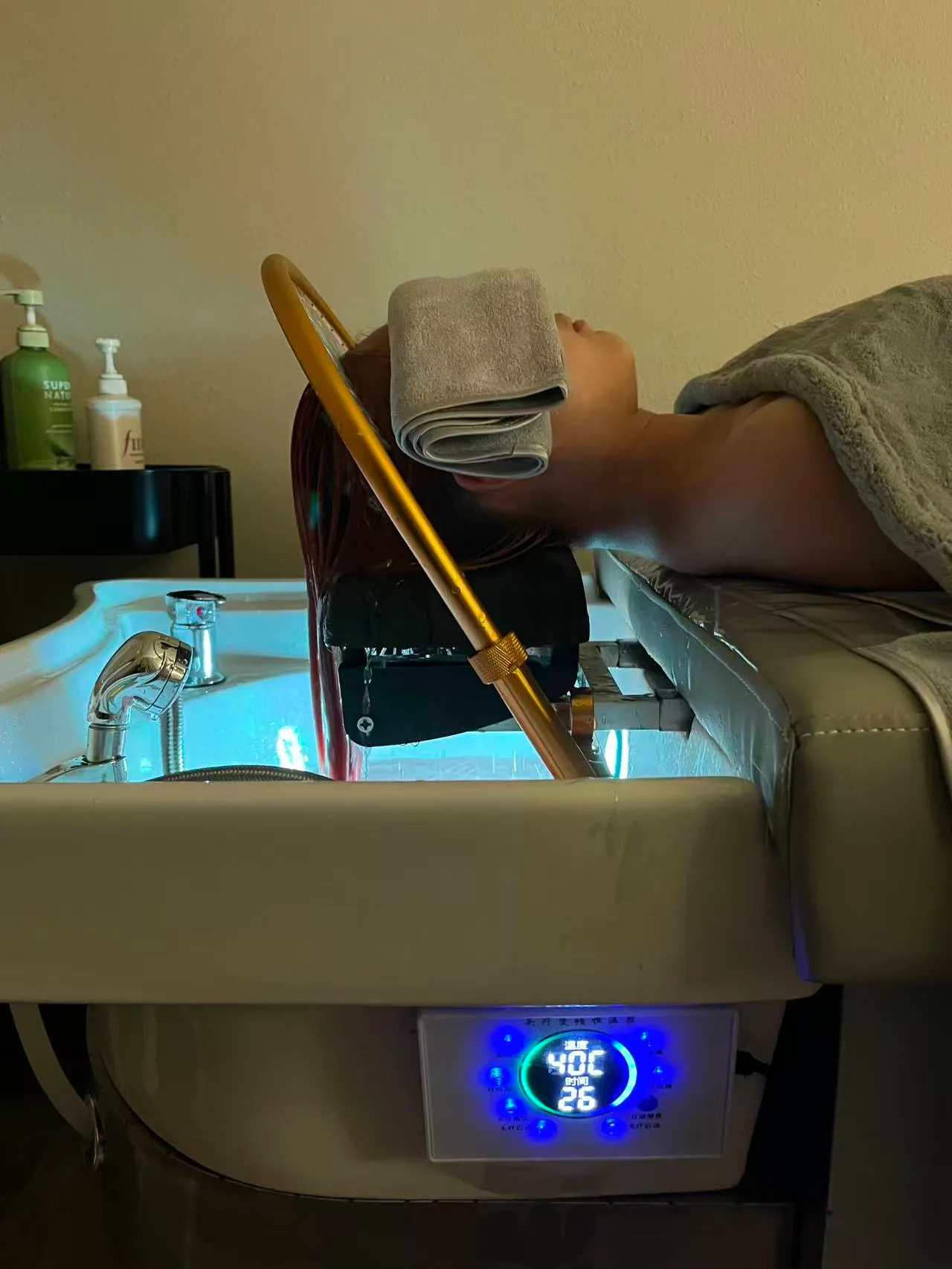 Side view of a client reclined at an O2 head spa basin with a towel over the eyes, a gold rinse arch over the hair and a blue digital water-temperature display on the basin