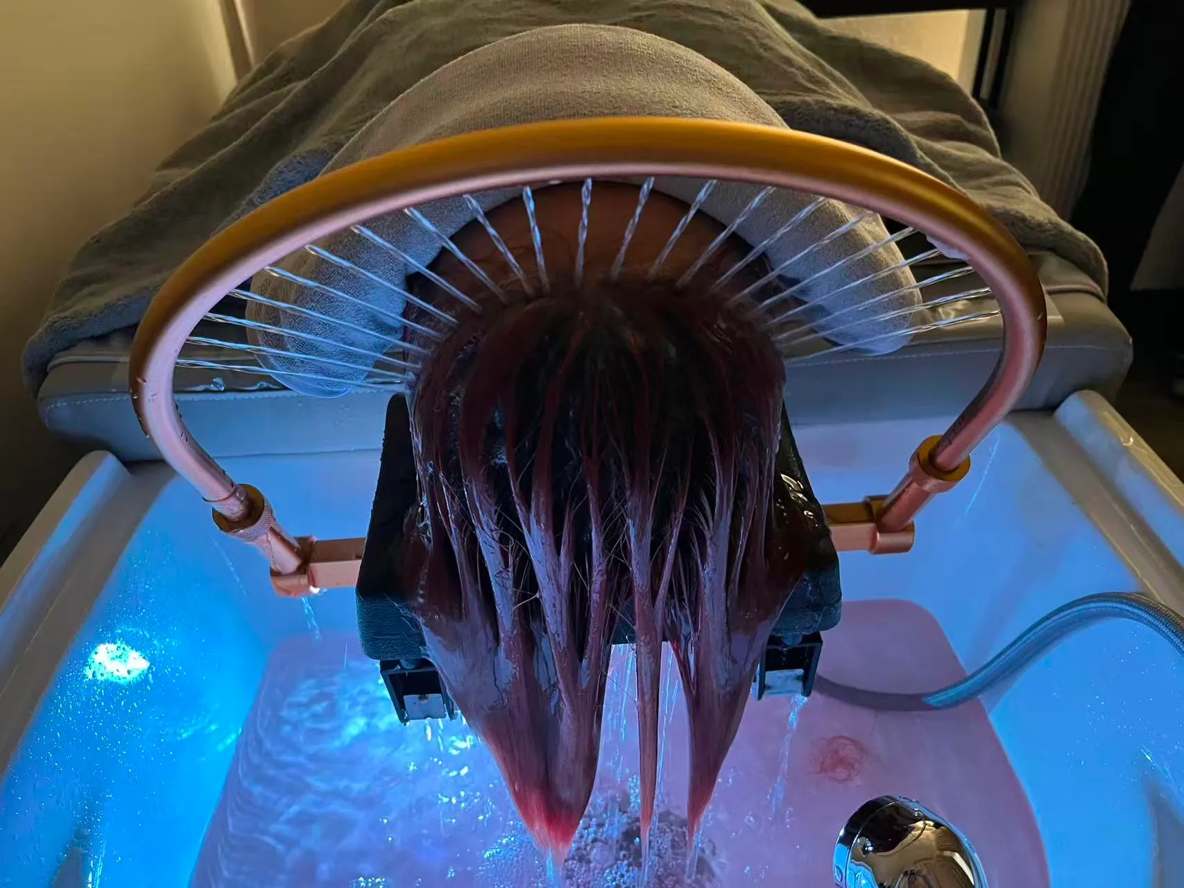 Close-up of a client's hair fanned through the gold waterfall arch over a blue-lit basin during waterfall hydrotherapy at O2 Head & Beauty Spa