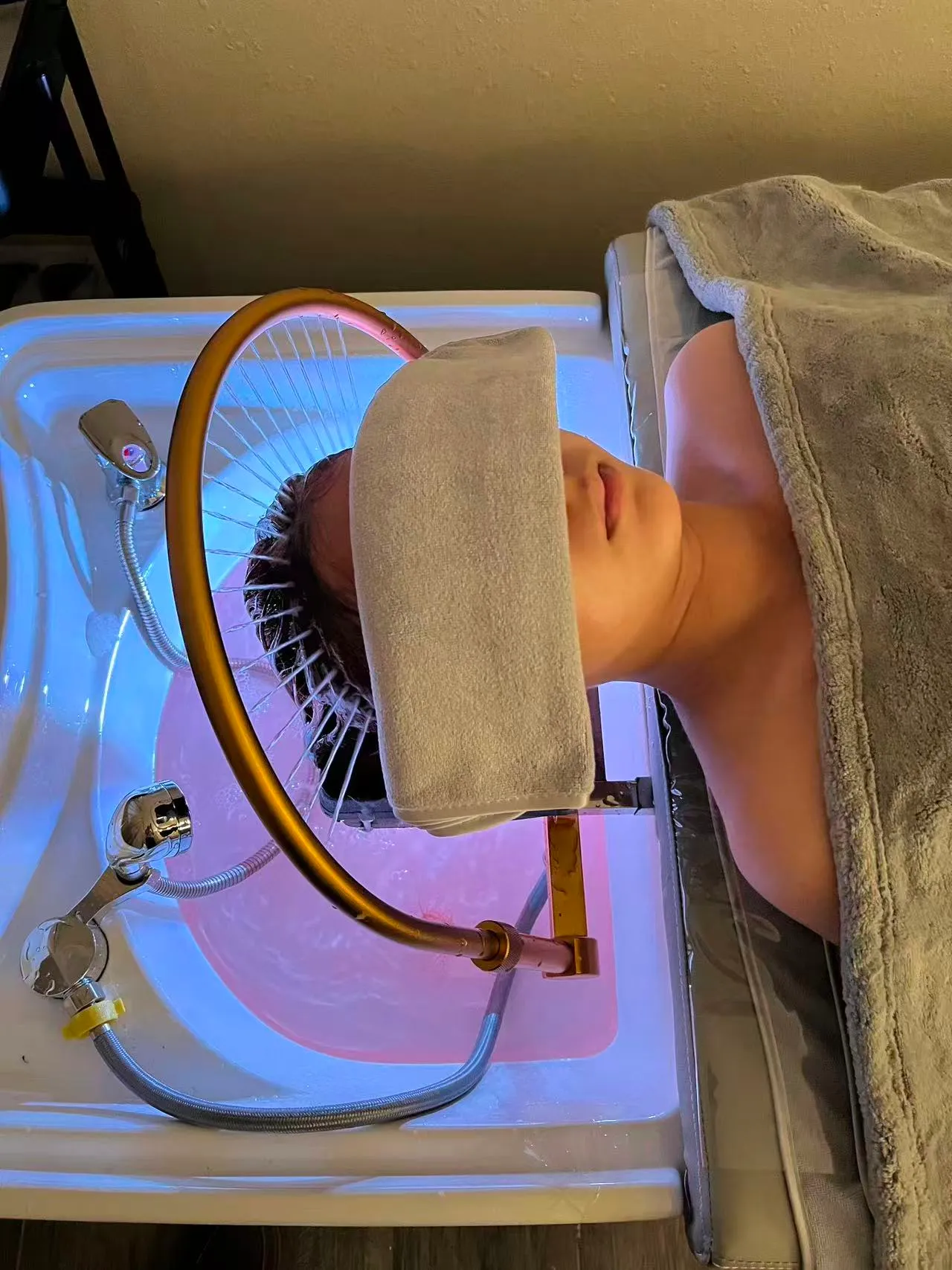 Client lying back at a head spa basin under the gold waterfall arch, a towel folded over the eyes